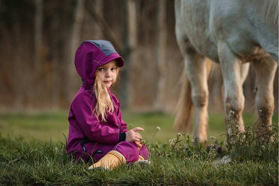 A little girl in a pink hooded jacket sits in the grass next to a white pony